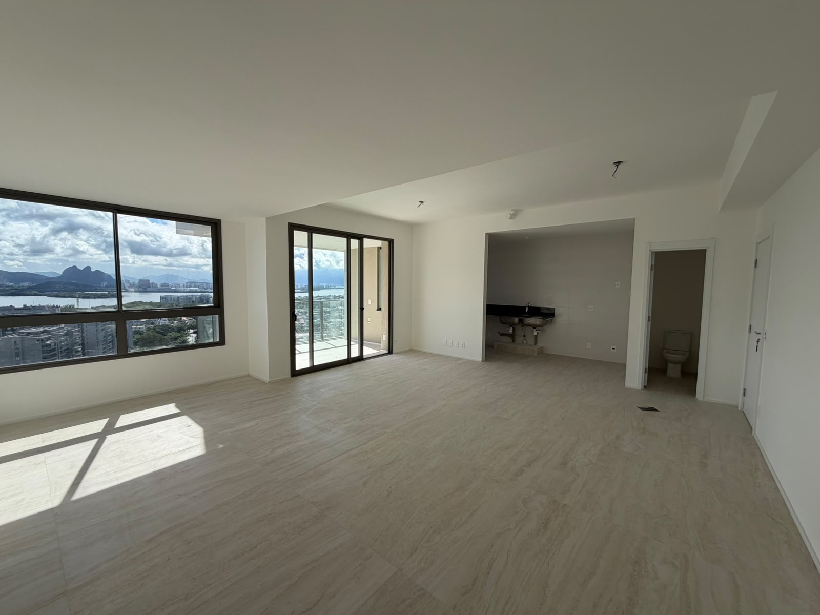 Sala ampla do apartamento no Atlântico Golf Absolut Design, na Barra da Tijuca, com piso claro, varanda e vista para a lagoa e os morros